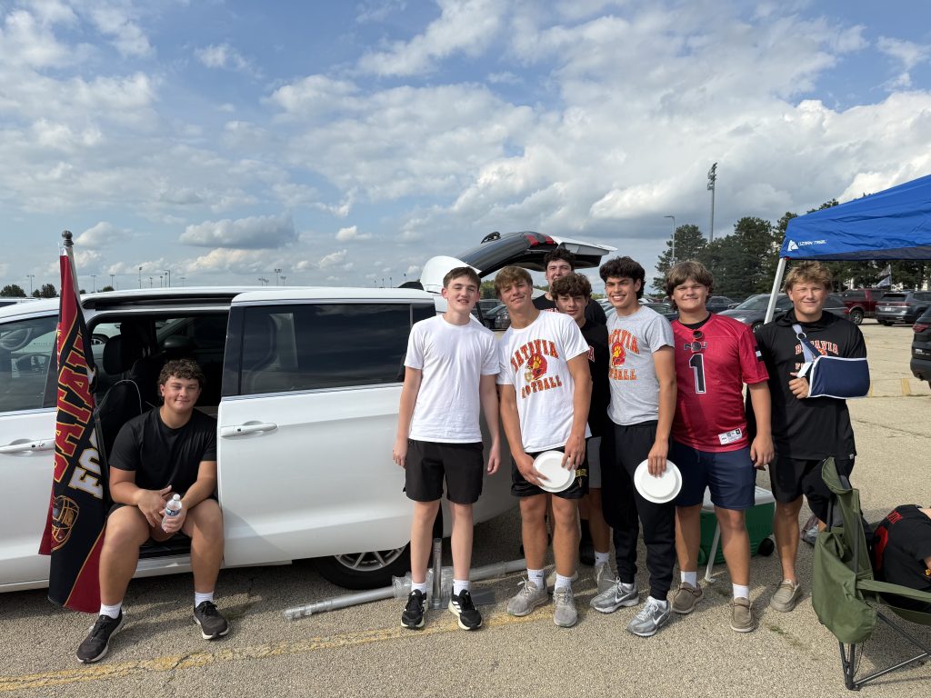 Group of high school football players posing next to a white van with a custom 3x5 team flag mounted on the vehicle. The team is dressed in athletic gear, and the setting appears to be a parking lot before a game or event.